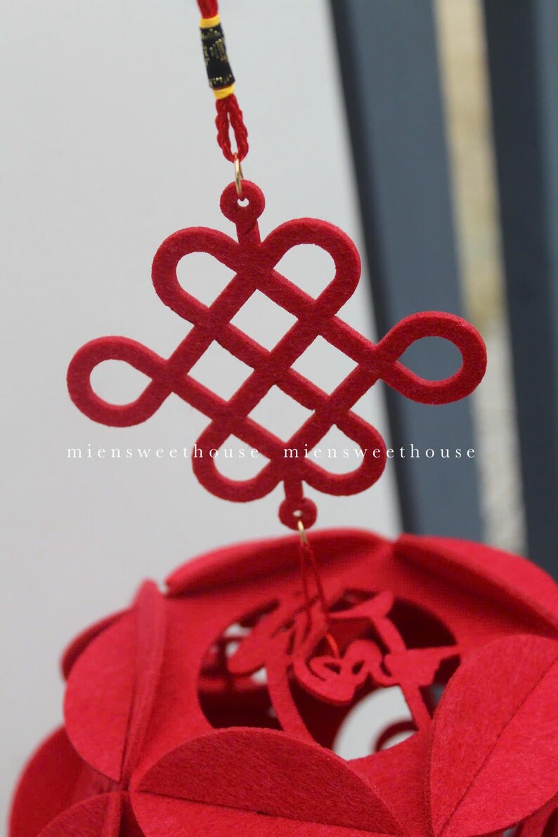 May include: A red felt Chinese knot, also known as a lucky knot, hangs from a red string. The knot is a symbol of good fortune and is often used in Chinese decorations. The knot is attached to a red felt ball with cutouts.