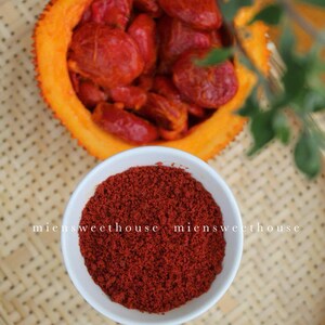 Pu&ograve; includere: Una ciotola bianca piena di polvere rosso vivo, probabilmente polvere di peperoncino, si trova su un tappetino di bamb&ugrave; intrecciato. Un frutto arancione tagliato a met&agrave; con semi rossi &egrave; visibile sullo sfondo.