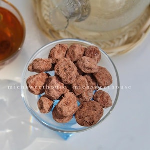 May include: A glass bowl filled with small, round, dark brown chocolate candies. The candies are arranged in a cluster and appear to be coated in a thin layer of cocoa powder.