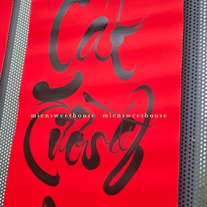 May include: A red scroll featuring black calligraphy. The text reads "Cat Cuong An" and includes the website "miensweethouse". The scroll is suspended by a bamboo rod with black detailing. A perforated metal panel forms the background.