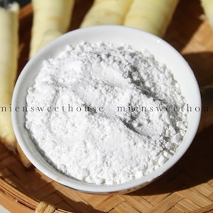 May include: A white bowl filled with white powder, possibly starch or flour, sits on a woven bamboo tray. Several peeled, light yellow root vegetables are arranged around the bowl, suggesting a culinary theme.