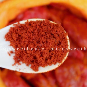 Pu&ograve; includere: Un cucchiaio di ceramica bianca pieno di polvere di paprika rosso vivo. Il cucchiaio &egrave; appoggiato su una ciotola di frutta rossa e arancione.