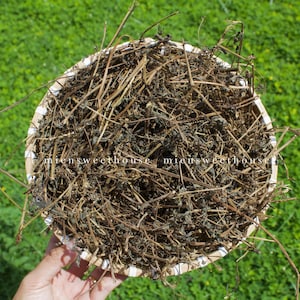 May include: A circular woven basket filled with dried herbs and stems. The herbs are a mix of brown and green, with a natural, organic appearance. The basket is held against a backdrop of green grass.