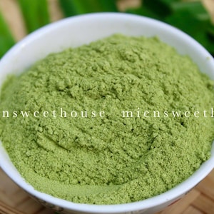 May include: A white bowl filled with a bright green powder. The powder is a vibrant green colour and has a fine texture. The bowl is sitting on a woven surface.