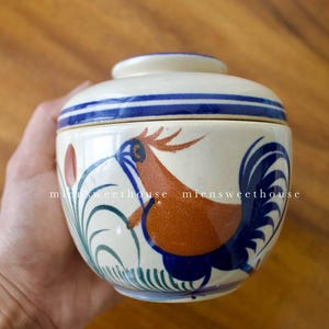 May include: A white ceramic container with a blue and green rooster design. The container has a lid and is hand-painted.