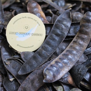 May include: A pile of dark brown, dried locust bean pods with a round, cream-colored sticker placed on top. The sticker features a heart icon, the text "MIEN SWEET HOUSE", "vietnamese herb, spices and more", and "@miensweethouse" in black.