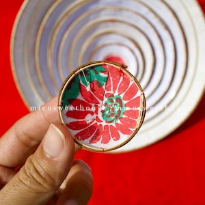 May include: A round, handmade ornament with a red and green floral design. The ornament is held in a hand against a red background. The text "miensweethouse" is visible on the ornament.