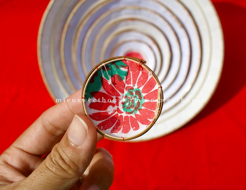 May include: A round, handmade ornament with a red and green floral design. The ornament is held in a hand against a red background. The text "miensweethouse" is visible on the ornament.