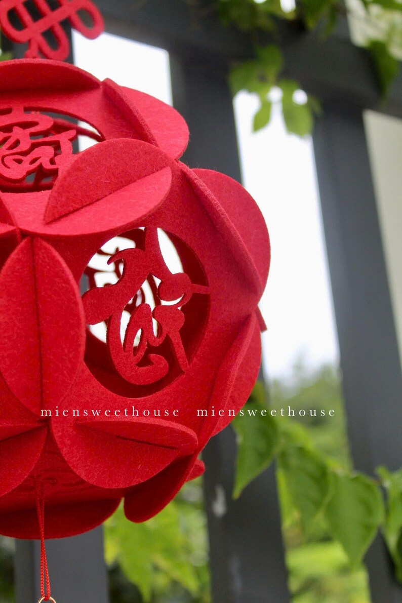 May include: A red felt Chinese lantern with intricate cutouts of Chinese characters. The lantern is hanging from a string and has a gold tassel.