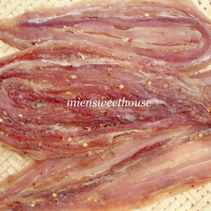 Op de afbeelding: Close-up van een gedroogd visproduct, gerold en in plakjes gesneden, met een roodachtig-roze kleur en een gevlekt uiterlijk. De vis ligt op een geweven bamboe oppervlak.