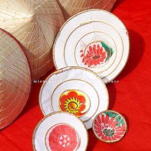 May include: A collection of conical straw hats and white, circular decorations with floral designs in red, yellow, and green. The items are arranged on a vibrant red surface.