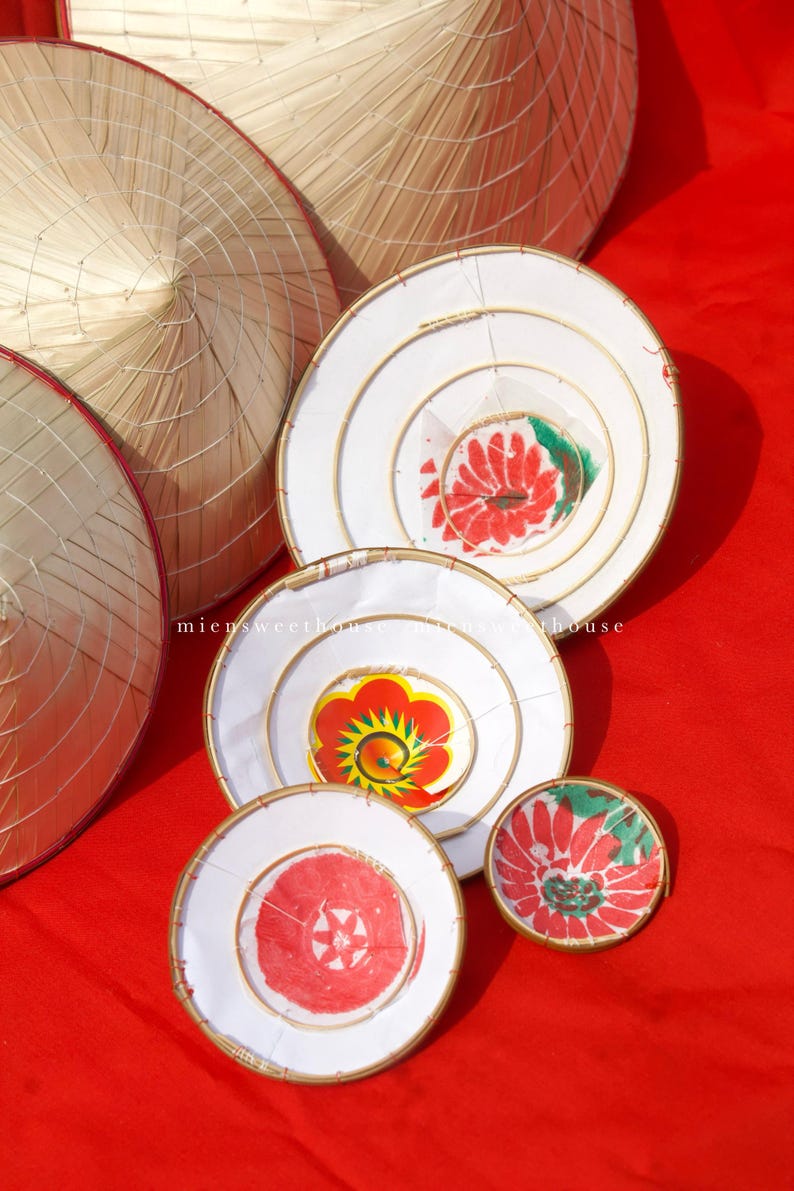 May include: A collection of conical straw hats and white, circular decorations with floral designs in red, yellow, and green. The items are arranged on a vibrant red surface.