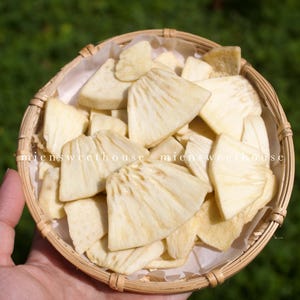 May include: A round woven basket filled with dried, light-yellow jackfruit slices. The basket is lined with white paper and held by a hand against a blurred green background. The text "miensweethouse" is visible on the basket.