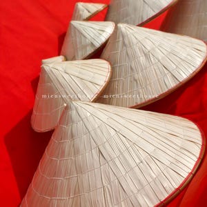 May include: A collection of traditional Asian conical hats made from woven natural fibers. The hats are light beige with a red trim, arranged on a vibrant red background. The text "miensweethouse" is visible on some hats.
