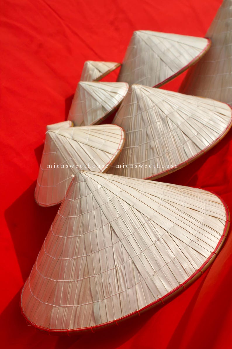 May include: A collection of traditional Asian conical hats made from woven natural fibers. The hats are light beige with a red trim, arranged on a vibrant red background. The text "miensweethouse" is visible on some hats.