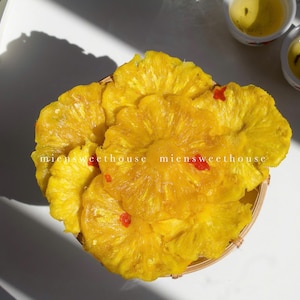 Op de afbeelding: Een close-up van een bord met gedroogde ananasreepjes, gerangschikt in een cirkelvorm. De ananasreepjes zijn felgeel en hebben een licht doorschijnende uitstraling. Er zijn kleine rode chilivlokken over de ananasreepjes gestrooid.