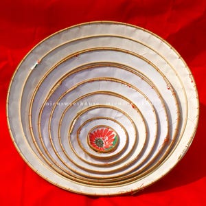 May include: A set of seven nested, circular woven trays with white fabric stretched across the interior. The trays are arranged in a spiral pattern, with a red and green floral design in the center. The background is a solid red.