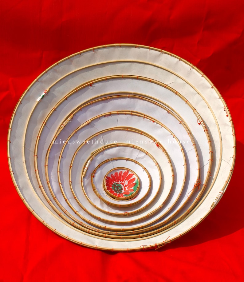 May include: A set of seven nested, circular woven trays with white fabric stretched across the interior. The trays are arranged in a spiral pattern, with a red and green floral design in the center. The background is a solid red.
