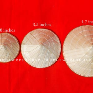 May include: Four conical straw hats of varying sizes, ranging from 1.9 to 4.7 inches in diameter, are displayed against a vibrant red background. The hats are made of woven straw and are a light beige color.