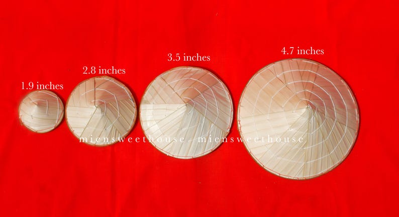 May include: Four conical straw hats of varying sizes, ranging from 1.9 to 4.7 inches in diameter, are displayed against a vibrant red background. The hats are made of woven straw and are a light beige color.