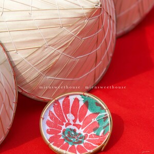 May include: Close-up of several conical hats made of woven bamboo and a small, round decorative item. The decorative item has a red floral design with green accents and a gold rim. The background is a solid red surface. The text "miensweethouse" is visible.