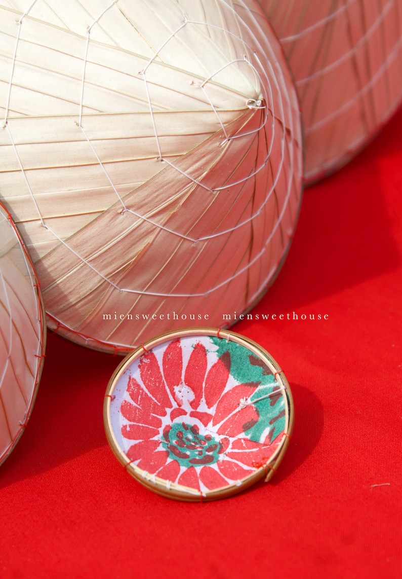 May include: Close-up of several conical hats made of woven bamboo and a small, round decorative item. The decorative item has a red floral design with green accents and a gold rim. The background is a solid red surface. The text "miensweethouse" is visible.