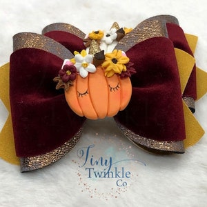 Pu&ograve; includere: Un fiocco di velluto bordeaux con bordino di glitter dorati e un fiocco di velluto giallo con bordino di glitter dorati. I fiocchi sono sormontati da una piccola zucca con un corno di unicorno e una corona di fiori.