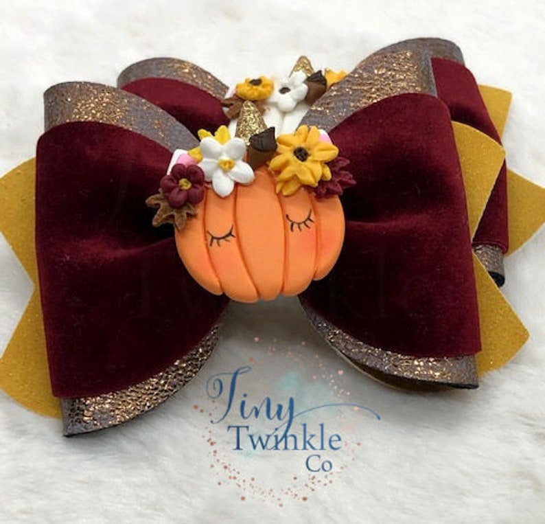 Pu&ograve; includere: Un fiocco di velluto bordeaux con bordino di glitter dorati e un fiocco di velluto giallo con bordino di glitter dorati. I fiocchi sono sormontati da una piccola zucca con un corno di unicorno e una corona di fiori.