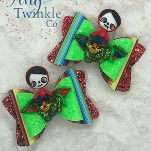 May include: Two colourful hair bows with a green, red and blue striped pattern. Each bow features a small, sculpted figure of a character from the movie Coco. The characters are wearing red and blue clothing and have a skull-like face.