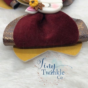Pu&ograve; includere: Un fiocco di velluto bordeaux con bordino dorato glitterato e un'applicazione di unicorno bianco con un dettaglio di fiore e foglia. Il fiocco &egrave; su una superficie bianca pelosa con il testo "Tiny Twinkle Co."