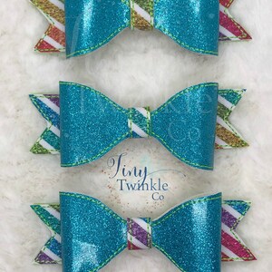 May include: Three turquoise glitter hair bows with white and multi-colored striped accents. The bows are arranged in a vertical stack. The bottom bow has the text "Tiny Twinkle Co" on the back.