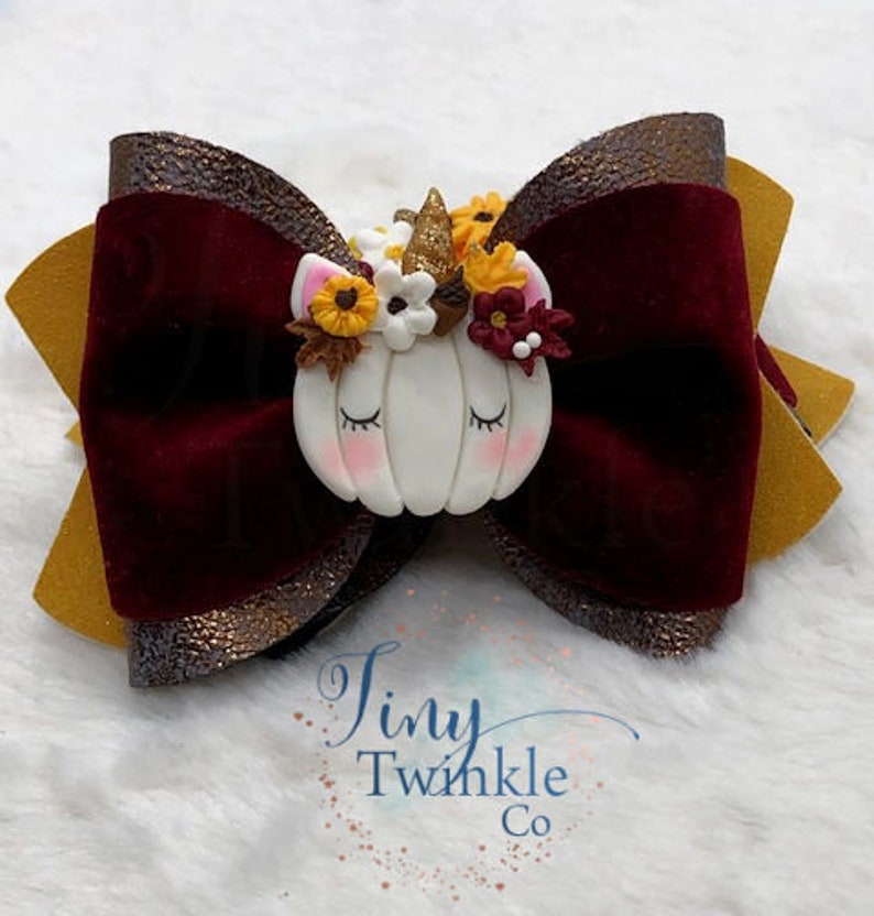 Pu&ograve; includere: Un fiocco di velluto bordeaux con accenti di glitter dorati e un unicorno bianco a forma di zucca con fiori autunnali. Il fiocco &egrave; decorato con il testo "Tiny Twinkle Co."