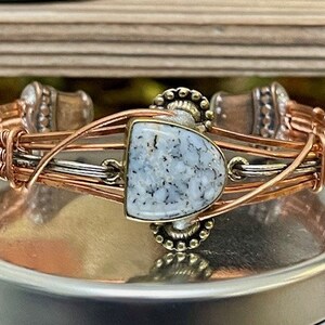 May include: A copper and silver wire wrapped bracelet with a white and gray stone set in the center. The stone is surrounded by decorative metal accents.