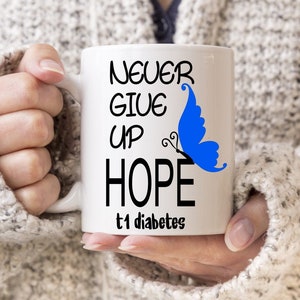 May include: A white ceramic mug featuring the text "NEVER GIVE UP HOPE t1 diabetes" in black, with a blue butterfly graphic. The mug is held by a person wearing a light grey knitted sweater.