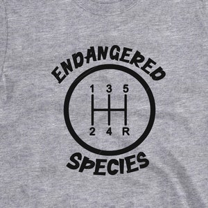 May include: Gray t-shirt with a black graphic of a car's gear shift pattern and the text "Endangered Species".