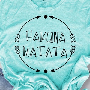 Könnte beinhalten: Ein türkisfarbenes T-Shirt mit einem schwarzen Kreisdesign mit dem Text "Hakuna Matata" und Pfeilen.