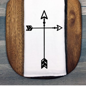 Puede incluir: Una toalla de cocina blanca con un diseño de flecha negra. La toalla está sobre una tabla de cortar de madera.