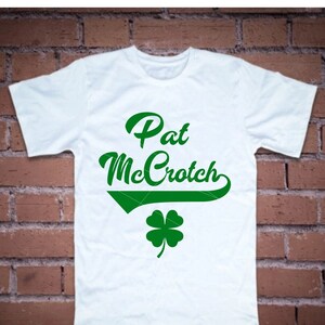 Op de afbeelding: Wit T-shirt met de tekst "Pat McCroch" in groene cursieve letters en een groen vierbladig klavertje.