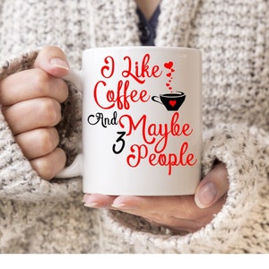 Puede incluir: Taza de café de cerámica blanca con texto negro que dice "Me gusta el café y tal vez 3 personas". El texto "tal vez" está en rojo. Hay pequeños corazones rojos sobre el texto "café" y una pequeña taza de café negra con un corazón rojo dentro.
