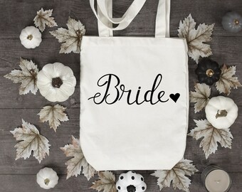 Op de afbeelding: Een witte draagtas met het woord "Bride" in zwart script en een zwart hart. De tas is omringd door witte en zwarte pompoenen, herfstbladeren en een kaars op een houten oppervlak.