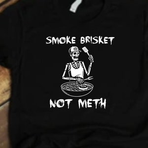 Può includere: Maglietta nera con una grafica bianca di uno scheletro che griglia un brisket. Il testo sulla maglietta recita "Smoke Brisket Not Meth".