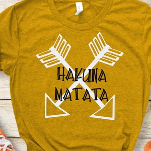 Könnte beinhalten: Ein senfgelbes T-Shirt mit einer weißen Grafik aus zwei gekreuzten Pfeilen mit dem Text "Hakuna Matata" in schwarzen Buchstaben.