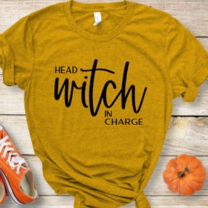 Op de afbeelding: Een mosterdgeel T-shirt met zwarte tekst die "Head Witch in Charge" luidt.