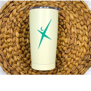 Puede incluir: Un vaso de acero inoxidable amarillo claro con un logotipo azul turquesa de una persona estilizada con una luna creciente sobre ella. El vaso está sobre una estera de paja tejida.