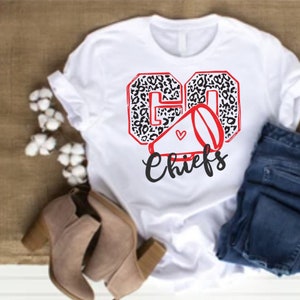 Puede incluir: Camiseta blanca con un diseño "GO Chiefs" en estampado de leopardo negro y rojo. El diseño incluye un megáfono rojo con un pequeño corazón rojo.