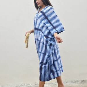 May include: A woman wearing a blue and white tie-dye maxi dress with a loose, flowing silhouette. She is walking on a concrete surface and holding a straw bag.