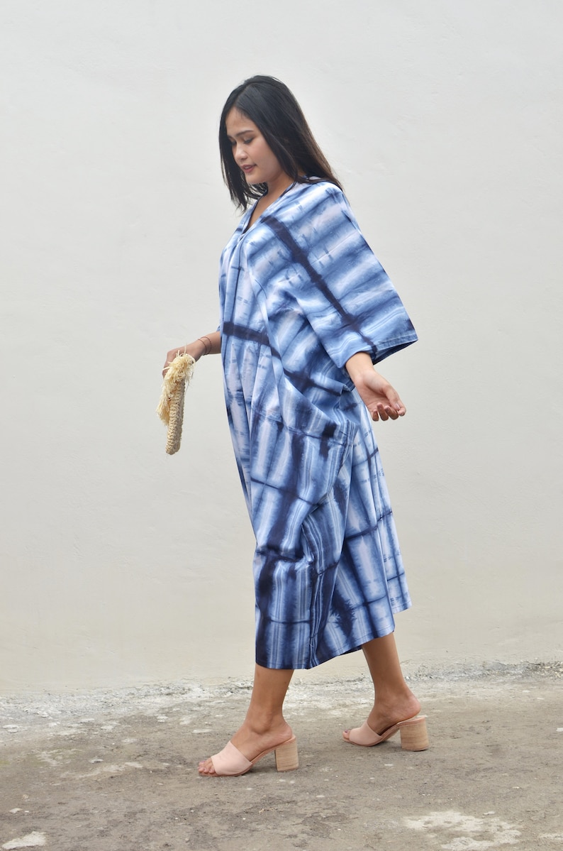 May include: A woman wearing a blue and white tie-dye maxi dress with a loose, flowing silhouette. She is walking on a concrete surface and holding a straw bag.
