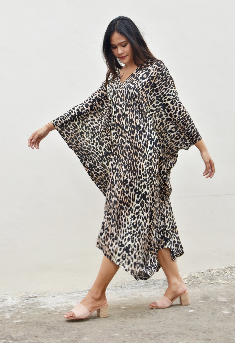 May include: A woman wearing a long, flowing leopard print kaftan dress with a V-neckline. She is walking on a concrete surface and wearing light pink heeled sandals.