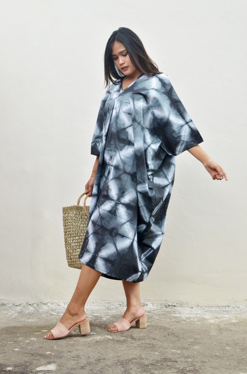 Puede incluir: Una mujer lleva un vestido maxi de color negro y blanco con estampado tie-dye, cuello en V y mangas cortas. Lleva un bolso de paja tejido y sandalias de tac&oacute;n nude.
