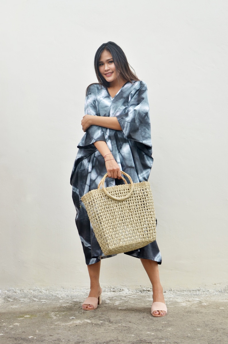 Puede incluir: Una mujer con un vestido maxi de tie-dye en blanco y negro con un gran bolso de mano de paja trenzada con asas de madera. Est&aacute; de pie sobre una superficie de hormig&oacute;n frente a una pared blanca.